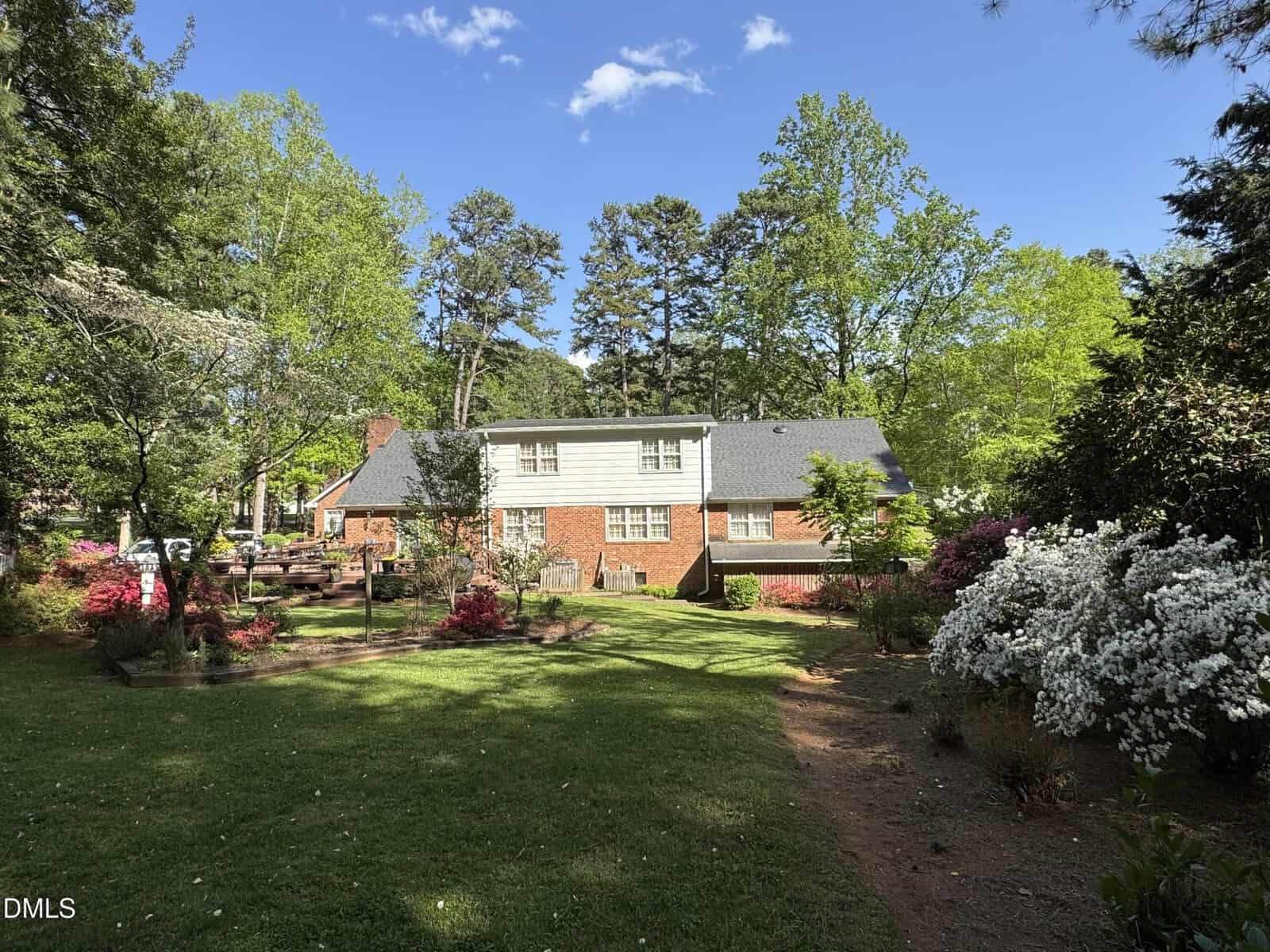 A two-story brick house with white siding sits in a lush, green yard with blooming flowers, tall trees, and a blue sky overhead. The scene is peaceful and sunlit.