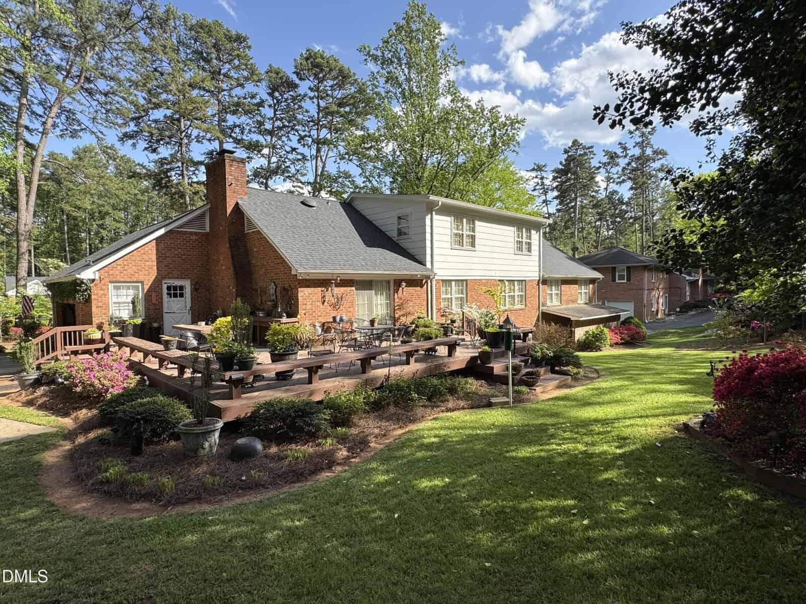 A brick house with a large wooden deck, surrounded by landscaped gardens, green lawn, and tall trees under a blue sky with scattered clouds.