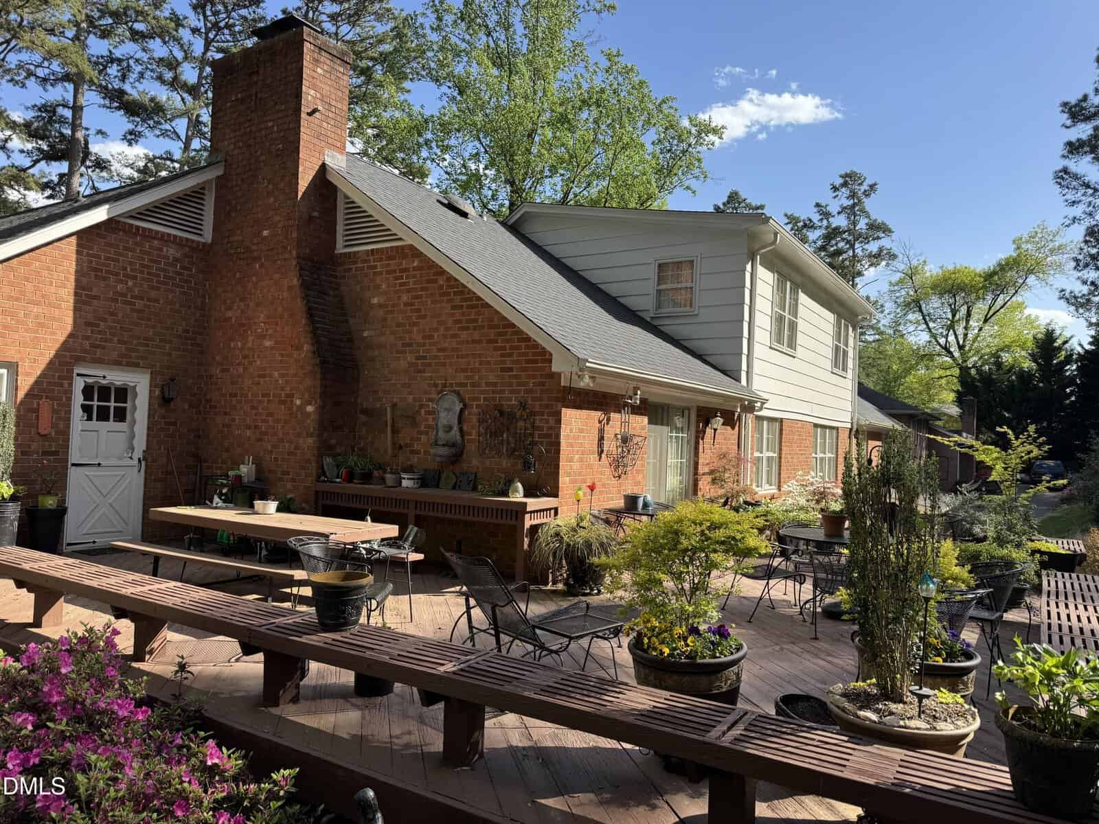 A spacious brick house with a large wooden deck, outdoor seating, potted plants, and greenery in the background on a sunny day. The deck features benches, tables, and decorative elements.