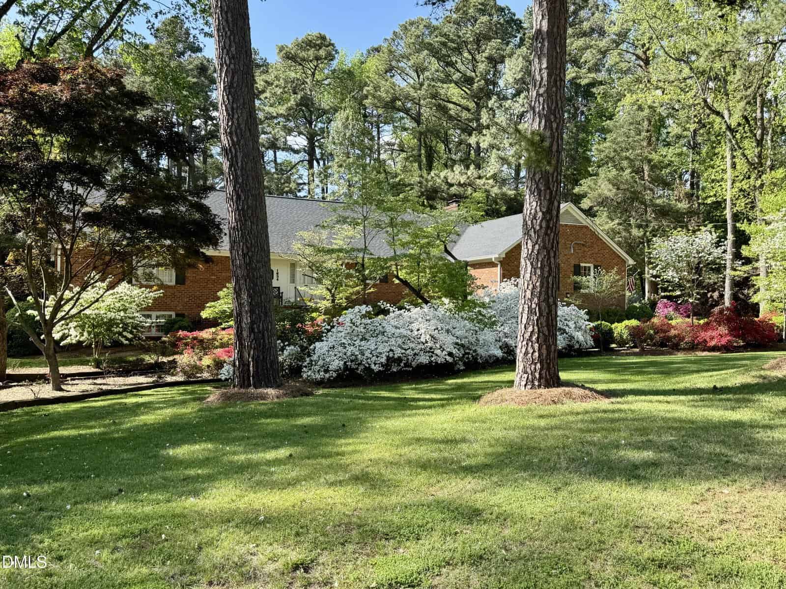 A brick house is surrounded by tall trees, green lawn, and flowering bushes, including azaleas and other shrubs, under a clear blue sky. Sunlight filters through the trees, creating a peaceful, lush garden scene.