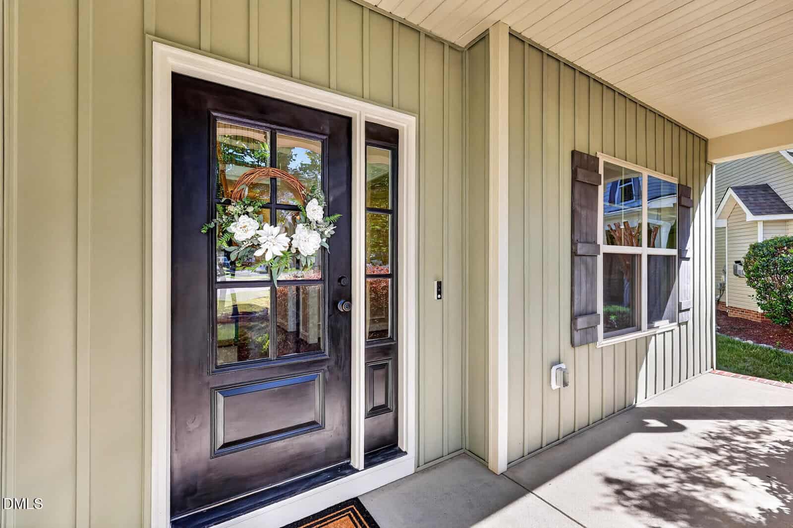 A front porch with beige vertical siding, a black door decorated with a white floral wreath, two large windows with dark shutters, and a concrete floor. Shrubs and grass are visible outside.