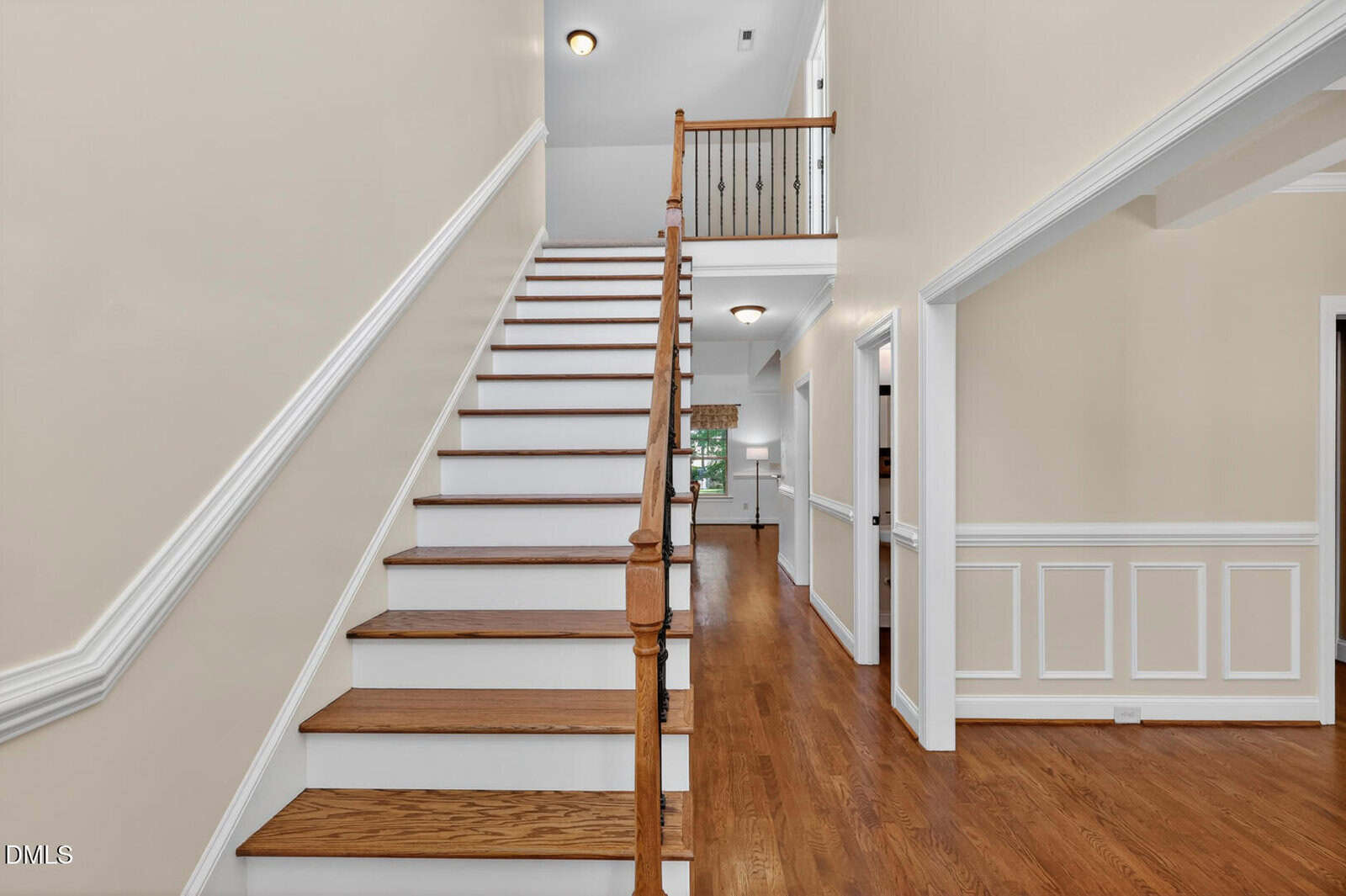 A bright foyer with hardwood floors features a wooden staircase with a white riser and a wooden handrail. The space has light beige walls, white trim, and leads to other rooms through open doorways.