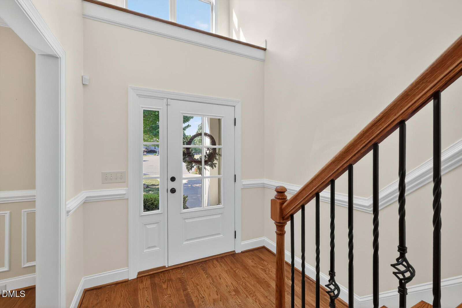 Bright entryway with a white front door featuring glass panels, wood trim, and side windows. Hardwood floors and a staircase with wooden handrail and black balusters are visible. Cream-colored walls and white wainscoting complete the space.