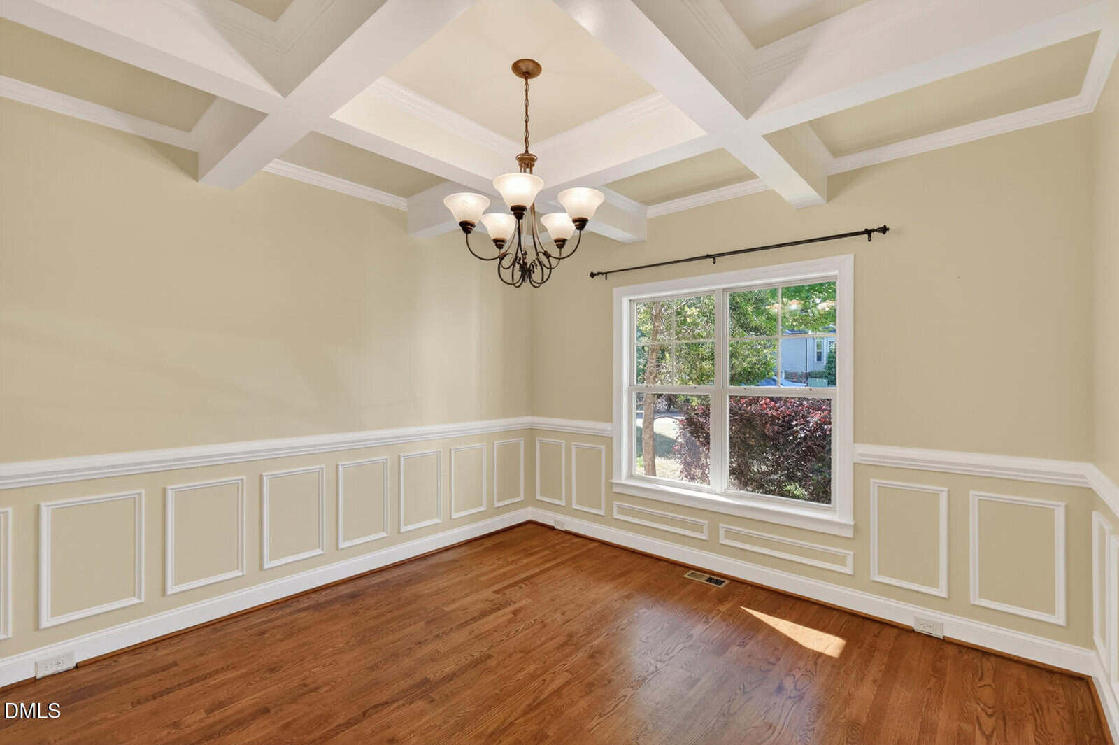 Bright, empty dining room with hardwood floors, cream-colored walls with white wainscoting, a large window, and a coffered ceiling featuring a bronze chandelier. Natural light enters through the window.