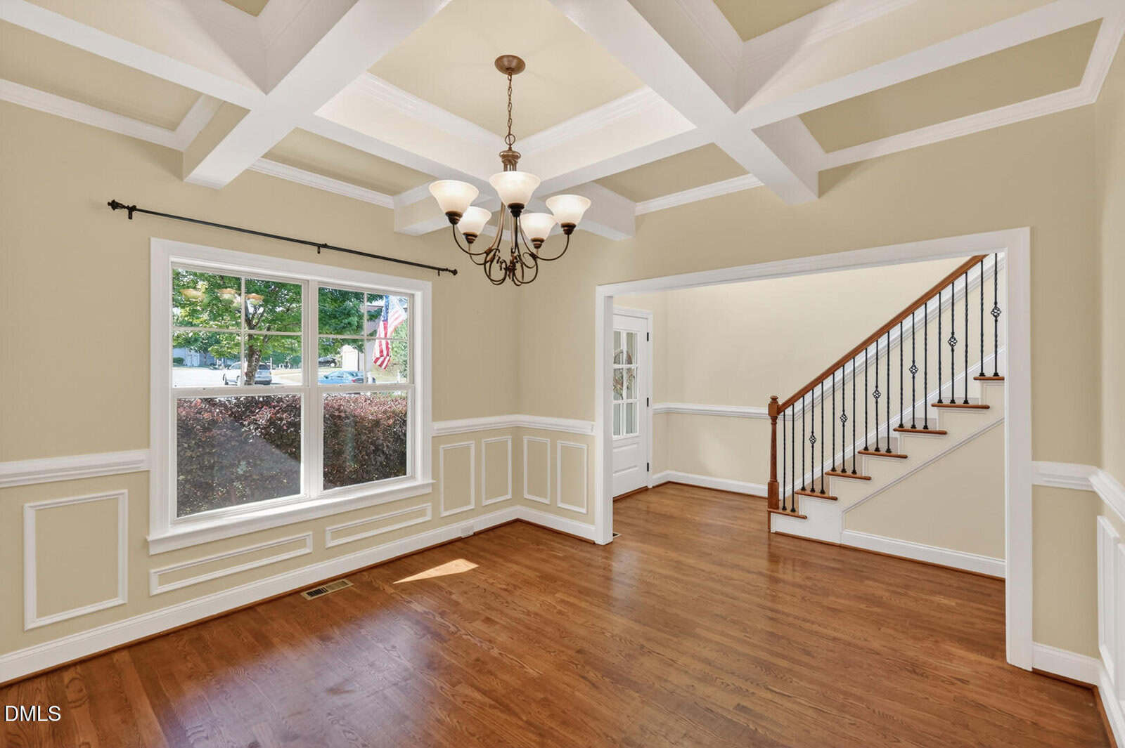 Bright, empty room with hardwood floors, a large window, coffered ceiling, chandelier, and white trim. An open doorway leads to a staircase with dark handrails and white risers.