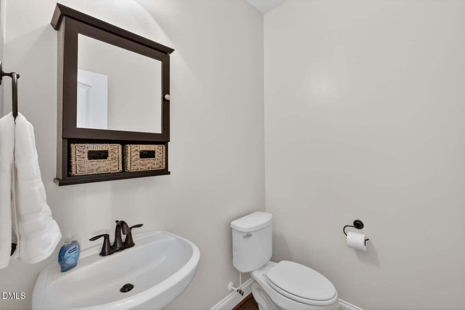 A small bathroom with a white sink, a soap dispenser, a mirror with two wicker baskets, a white toilet, a toilet paper holder, and a towel on a rack against a light beige wall.