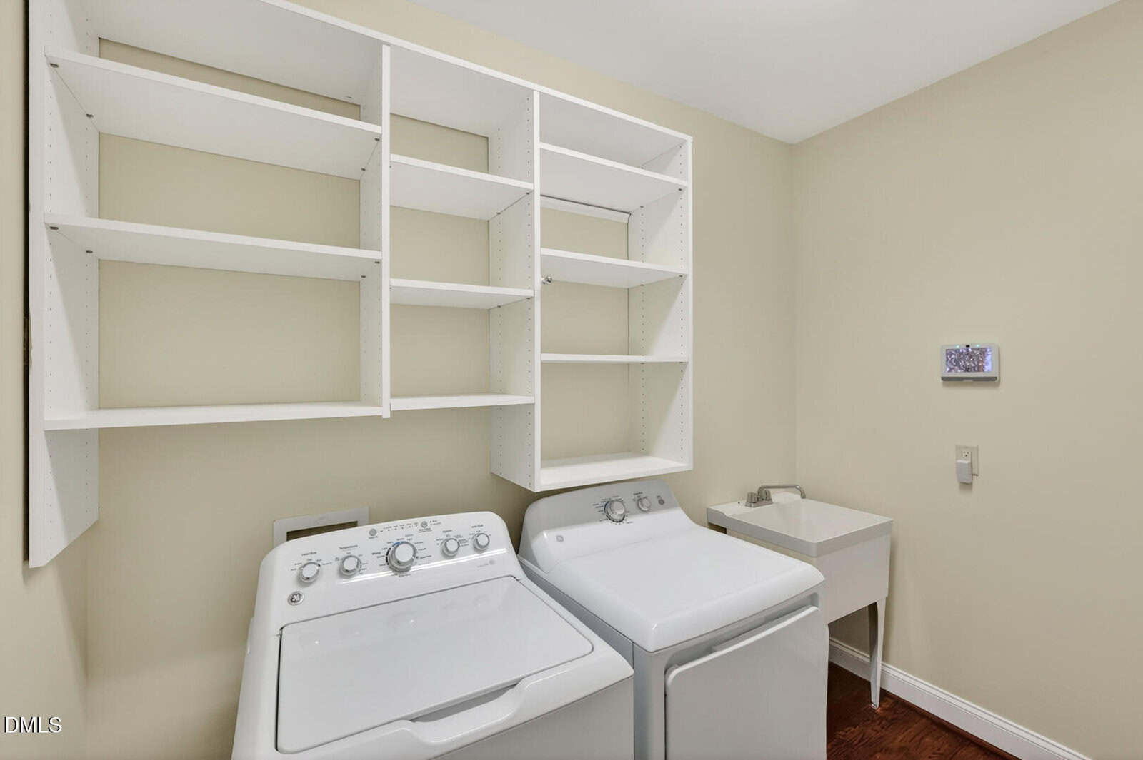 A laundry room with a white washing machine and dryer, open white shelves above, a small utility sink to the right, and beige walls.