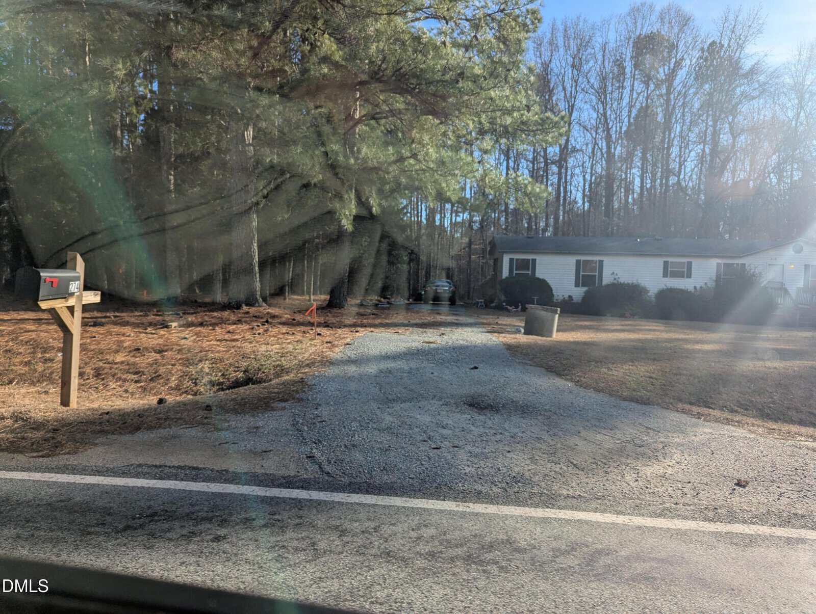 A gravel driveway leads to a single-story white house with green shutters surrounded by trees. A mailbox stands by the road, and sunlight filters through the trees casting shadows on the ground.