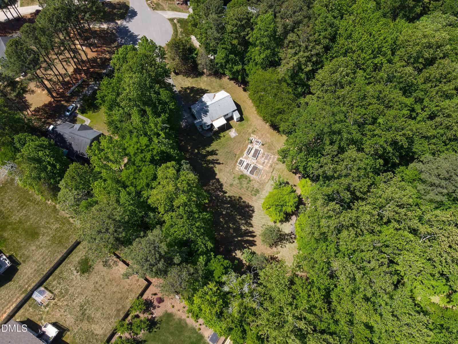 Aerial view of a house surrounded by dense trees, with a cleared yard containing garden beds and grassy areas, located in a residential neighborhood. Nearby houses and roads are partially visible among the greenery.