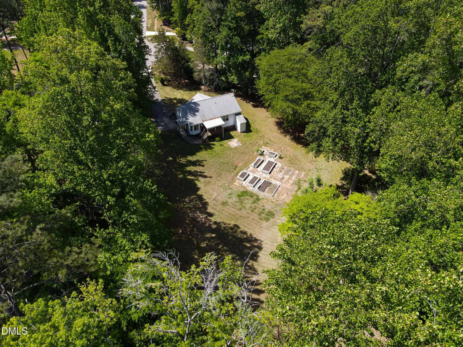 Aerial view of a small white house surrounded by trees, with several raised garden beds in a clearing in the backyard on a sunny day.