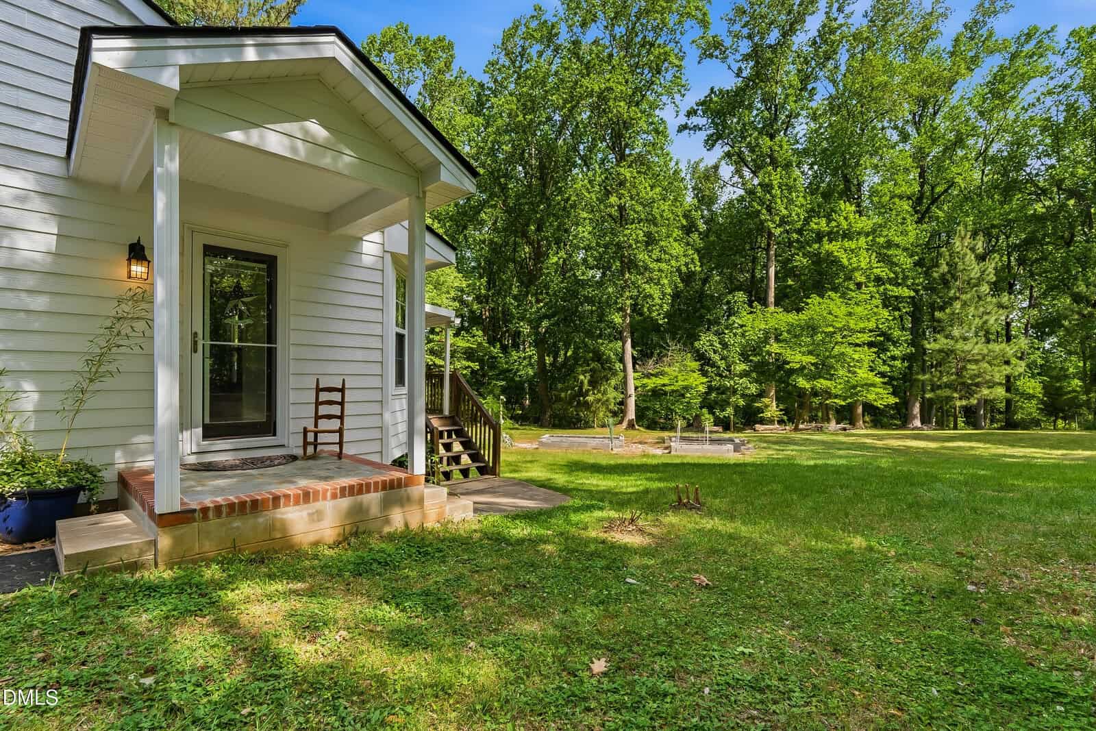 A white house with a small front porch, a wooden chair, and steps leading to a grassy yard bordered by lush green trees under a clear blue sky.