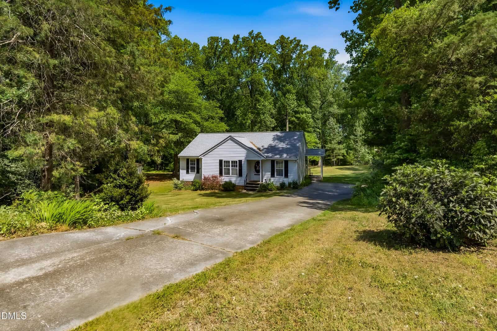 A small, single-story house with gray siding and white trim sits surrounded by lush green trees and bushes, with a long driveway leading up to it on a sunny day.