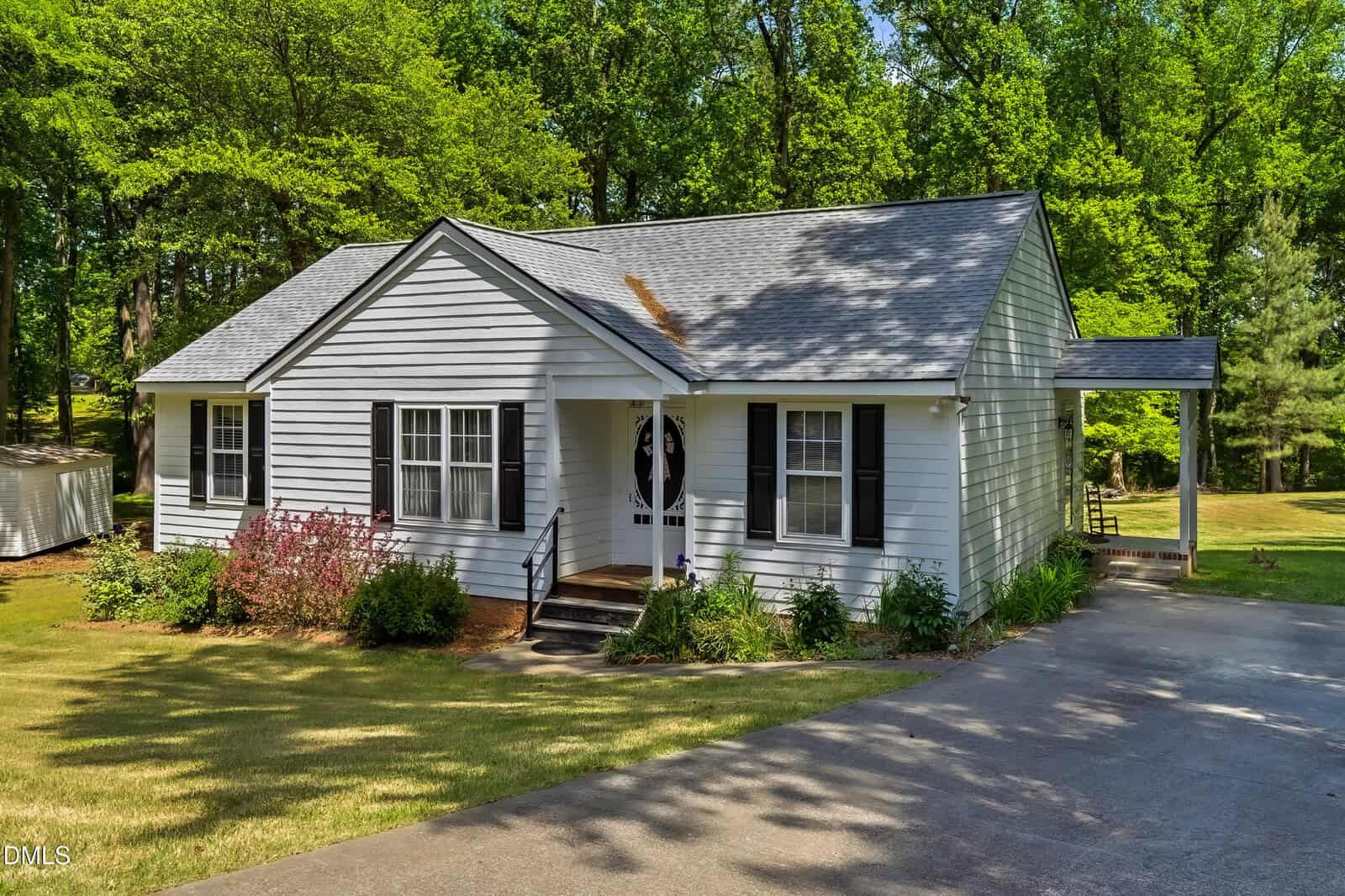 A small white house with black shutters, a front porch, and a carport sits on a neatly landscaped lawn surrounded by trees on a sunny day.