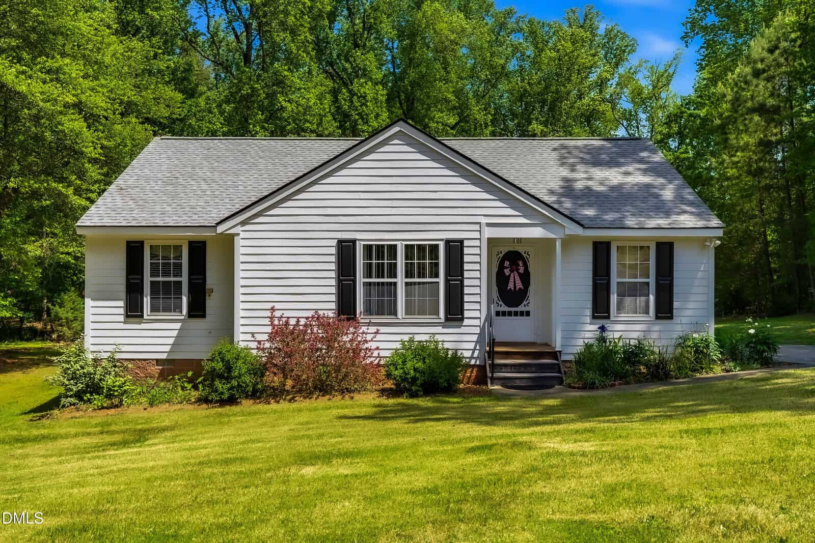 A small, white, single-story house with black shutters and a dark front door, surrounded by green grass, shrubs, and tall trees in the background under a clear blue sky.