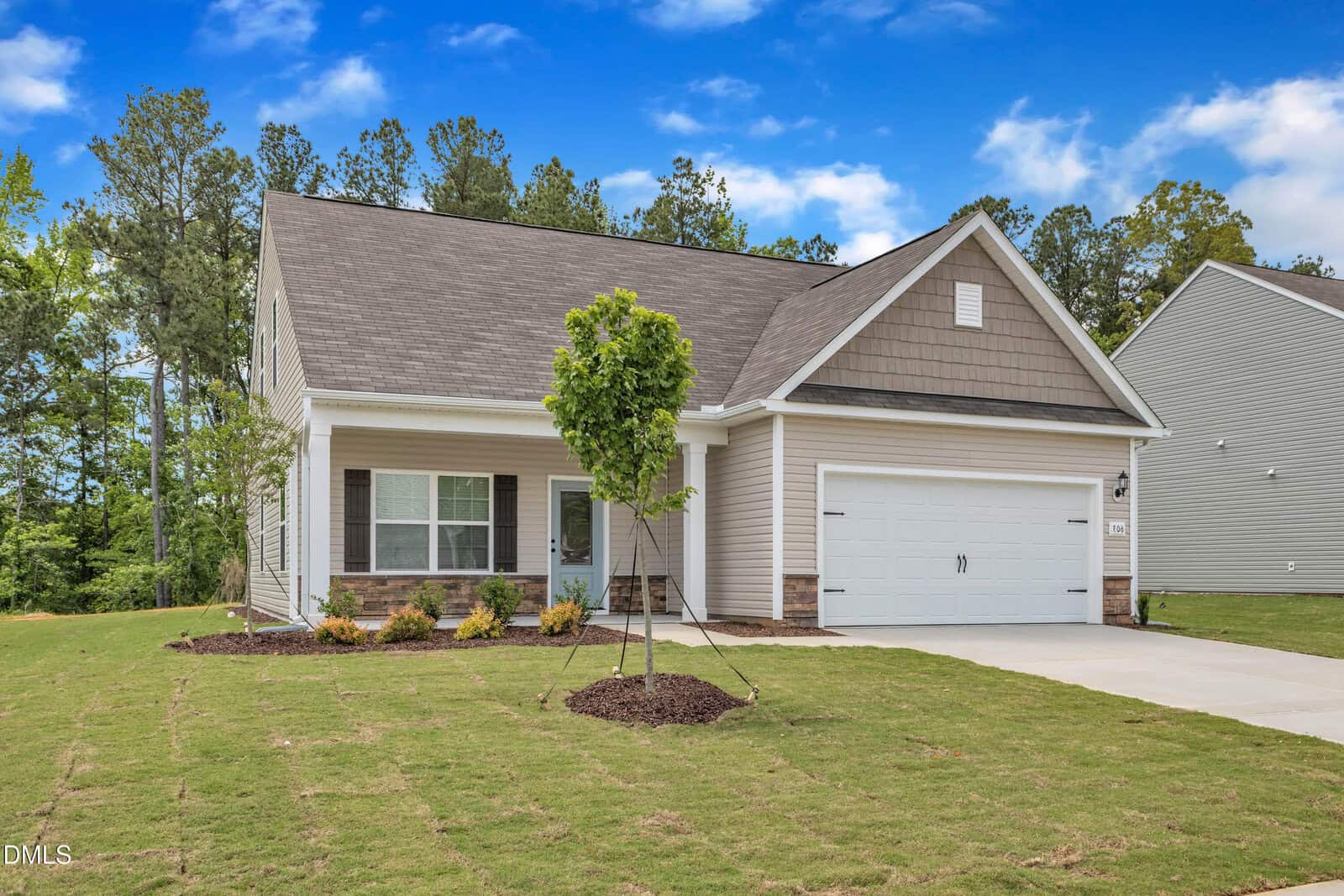 Single-story suburban house with beige siding, white trim, and a two-car garage. A small tree is planted in the front yard, and the lawn is freshly laid sod. Tall trees and a blue sky with clouds are in the background.