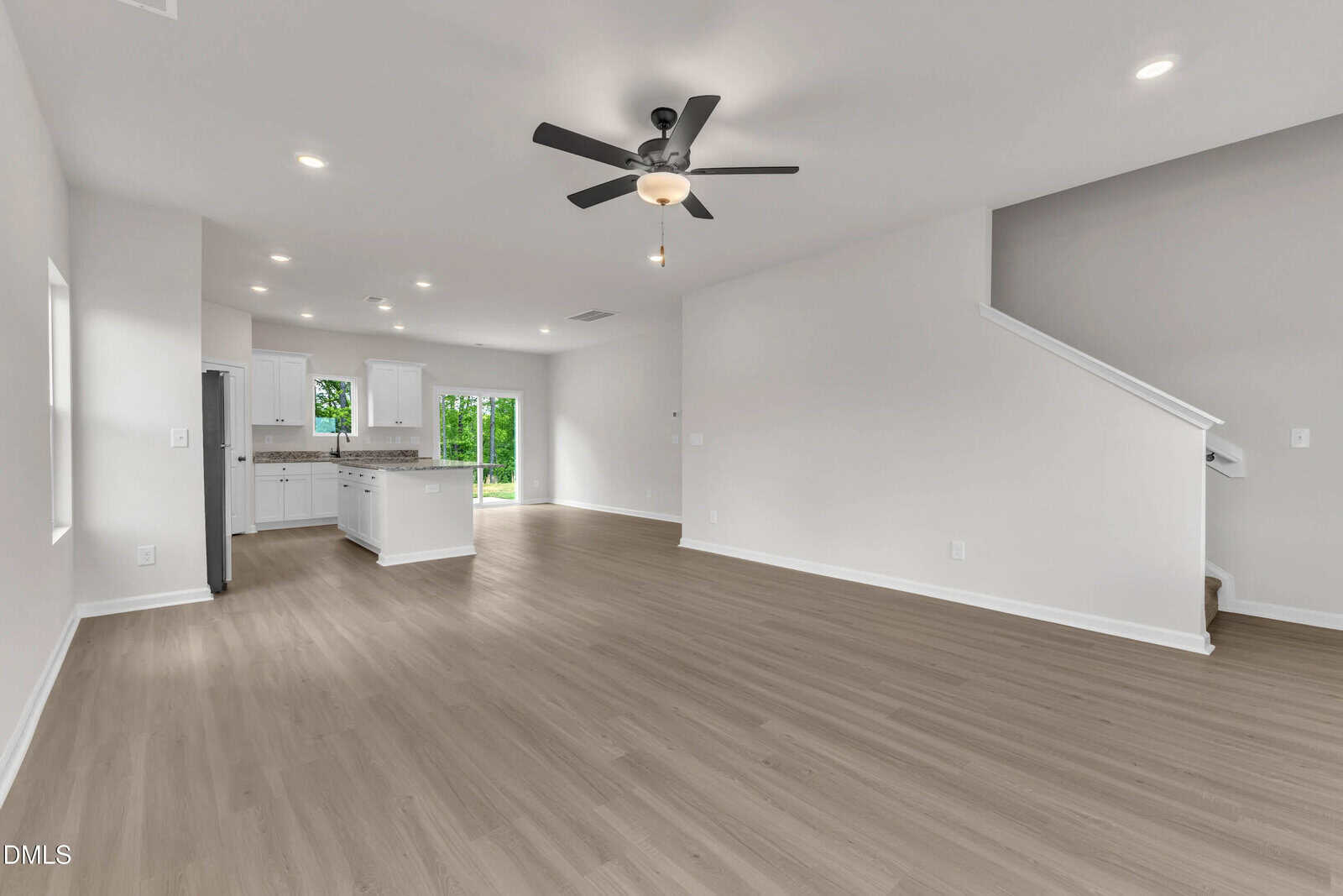 Spacious, modern open-concept living area with light wood flooring, white walls, recessed lighting, ceiling fan, kitchen with white cabinets and island, and a staircase leading upstairs. Large windows let in natural light.
