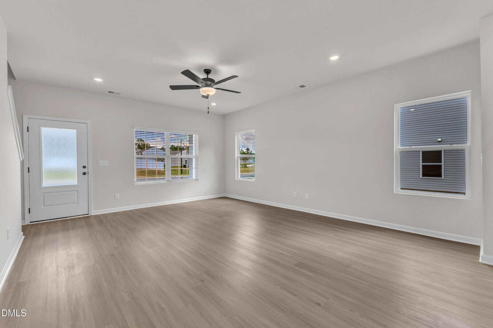 A spacious, empty living room with light wood floors, white walls, three windows, a ceiling fan with lights, and a white door with frosted glass panel. Natural light brightens the room.