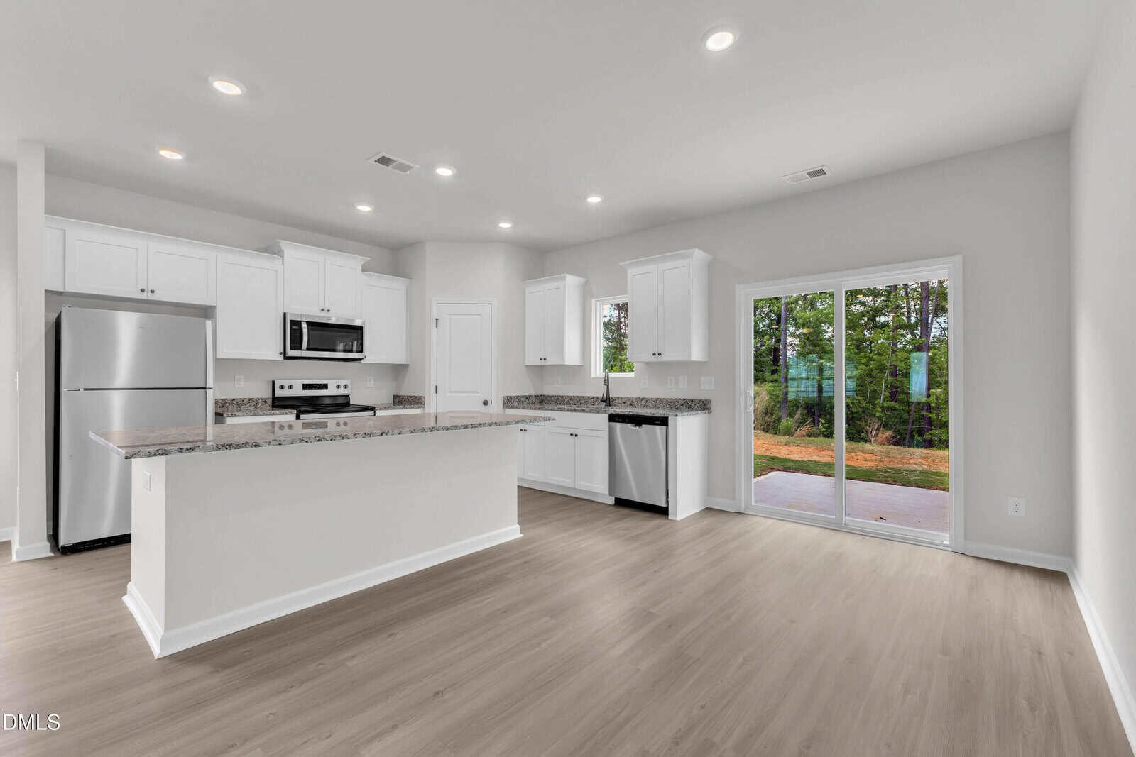 Modern kitchen with white cabinets, stainless steel appliances, granite countertops, and a large island. Light wood flooring and sliding glass doors open to a patio with a view of trees outside.