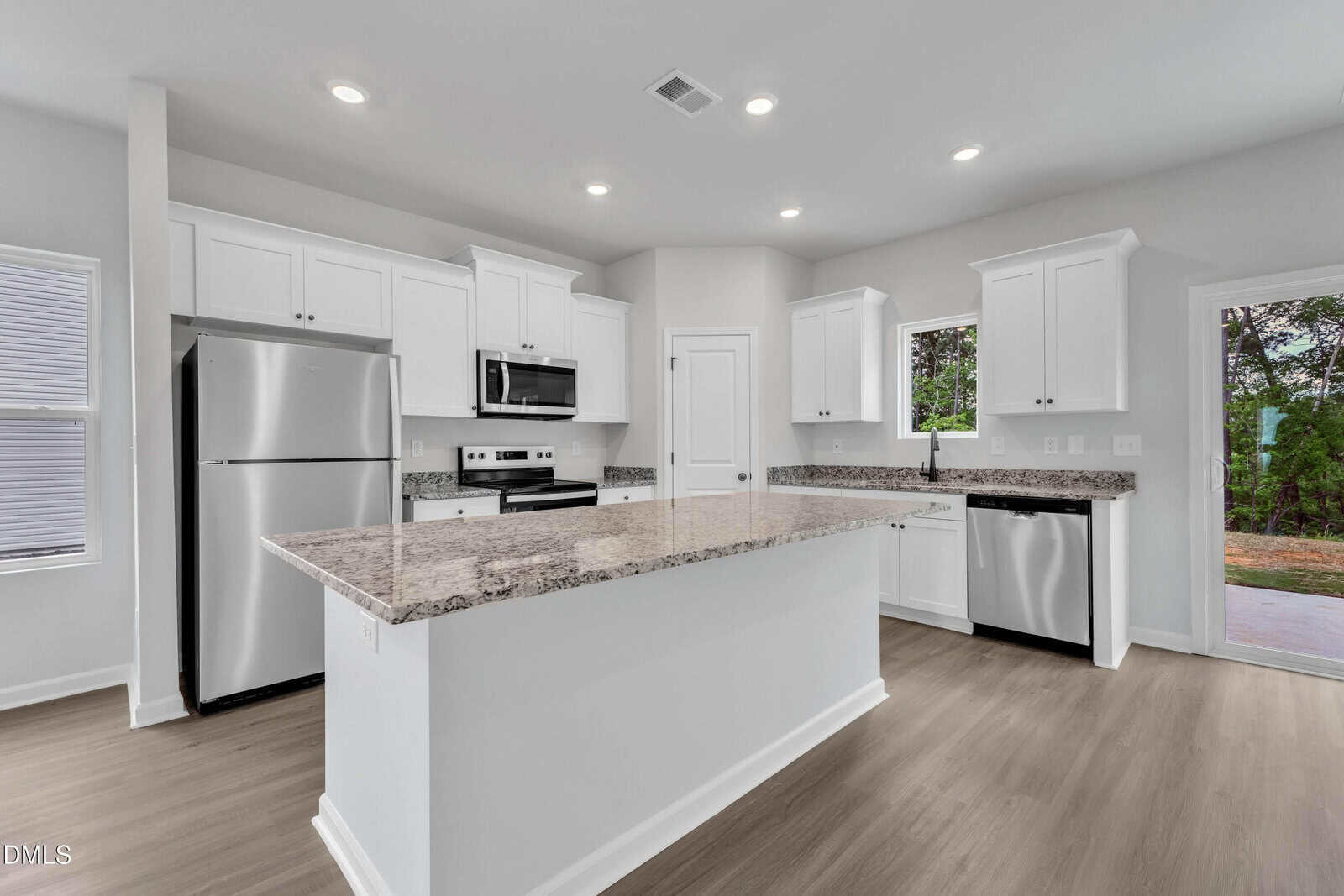 Modern kitchen with white cabinets, stainless steel appliances, granite countertops, a large center island, wood-look flooring, and windows providing natural light with views of trees and a patio outside.