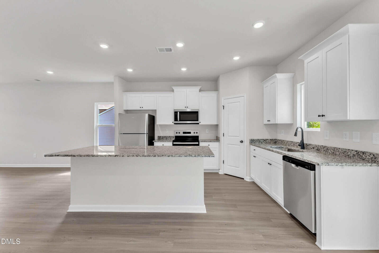 Modern kitchen with white cabinets, stainless steel appliances, granite countertops, light wood floors, and recessed ceiling lights. There is a large island, a dishwasher by the sink, and a pantry door to the right of the stove.
