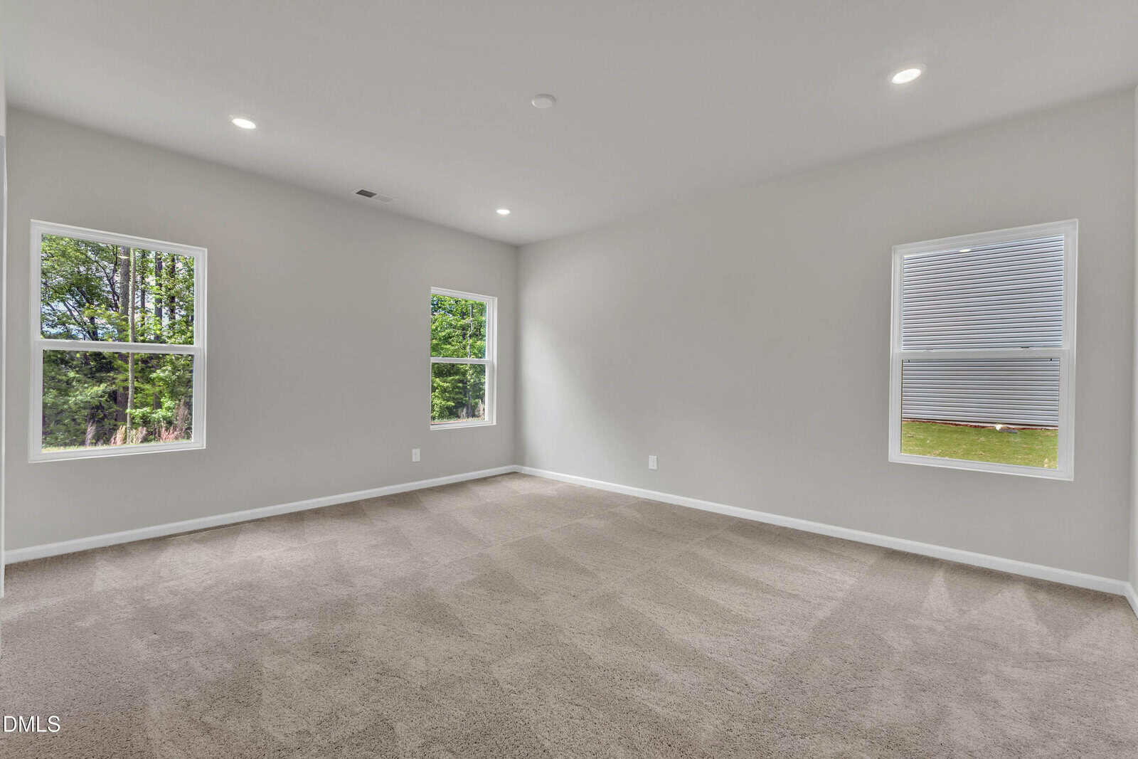 Empty room with beige carpet, light gray walls, and three windows. Two windows show green trees outside, and one window faces a neighboring building. The ceiling has recessed lighting.