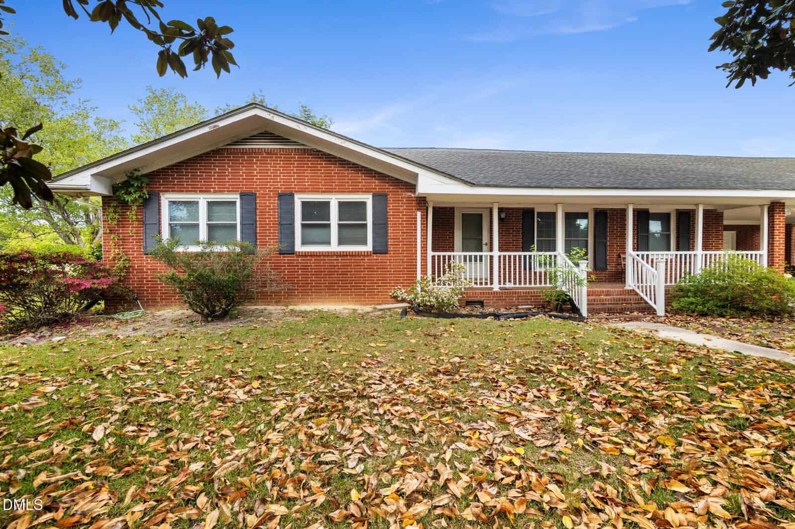 Single-story brick house with dark shutters, a covered front porch with white railings, and steps leading to the entrance. The lawn is scattered with fallen leaves, and bushes line the front of the house.