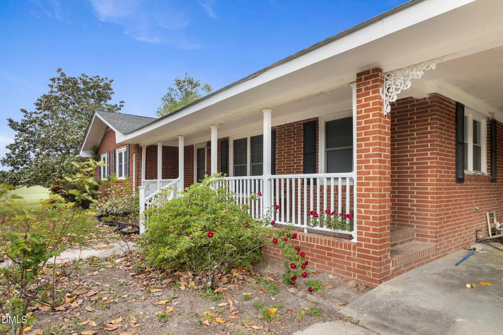 A red brick house with white trim and a covered front porch featuring white railings. Bushes with red flowers line the porch, and fallen leaves are scattered on the ground. The sky is clear and blue.