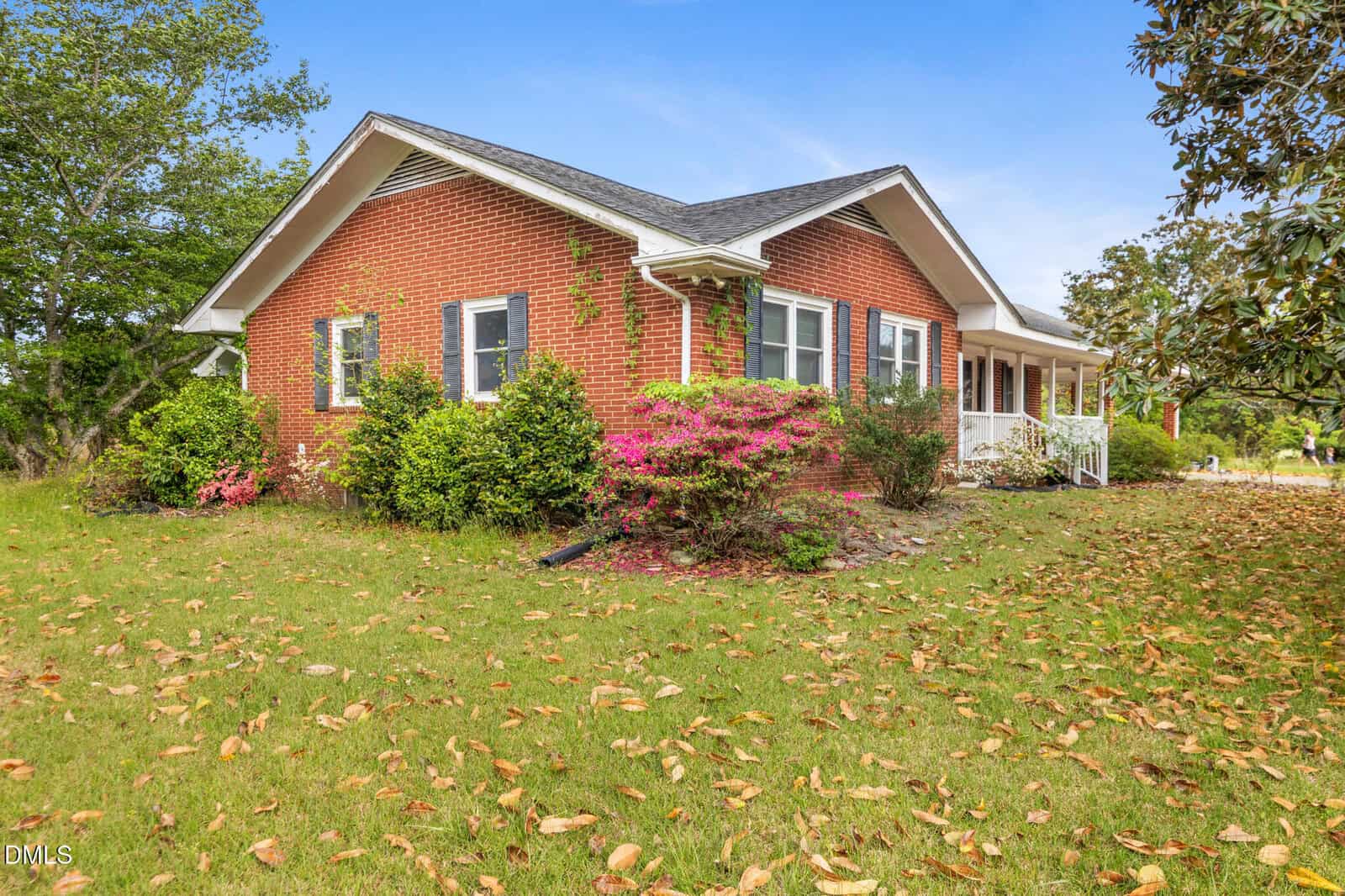 A single-story red brick house with white trim, surrounded by green shrubs and flowering bushes. The yard has scattered fallen leaves, and a white porch is visible along one side of the house. Blue sky is overhead.