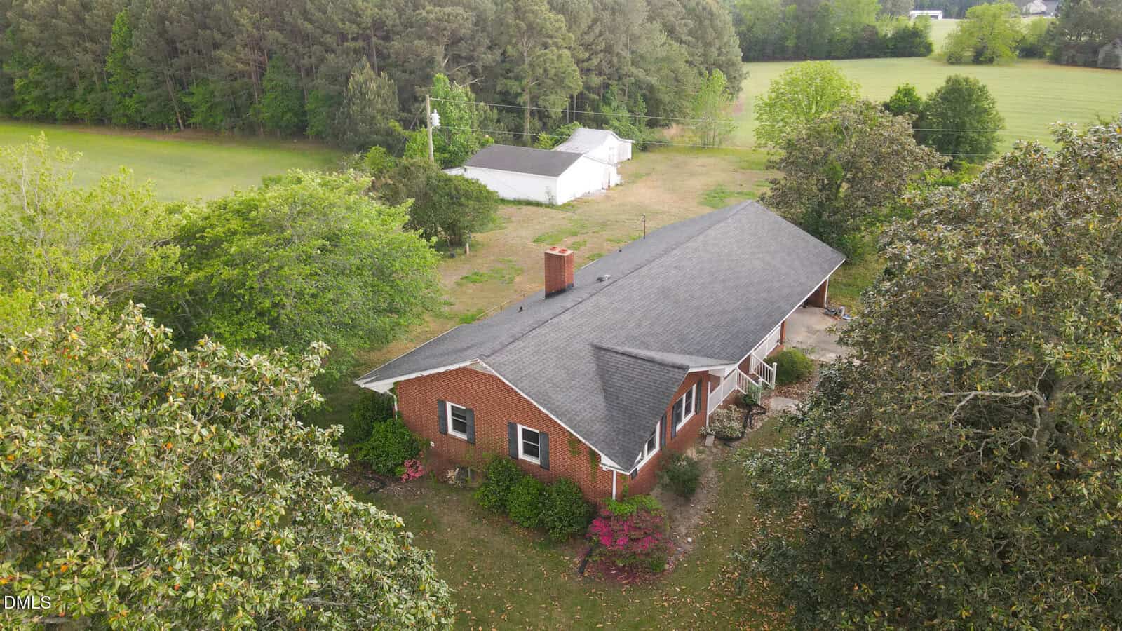 Aerial view of a red brick house with a dark roof, surrounded by green trees and shrubs, with a white outbuilding and open grassy area in the background.