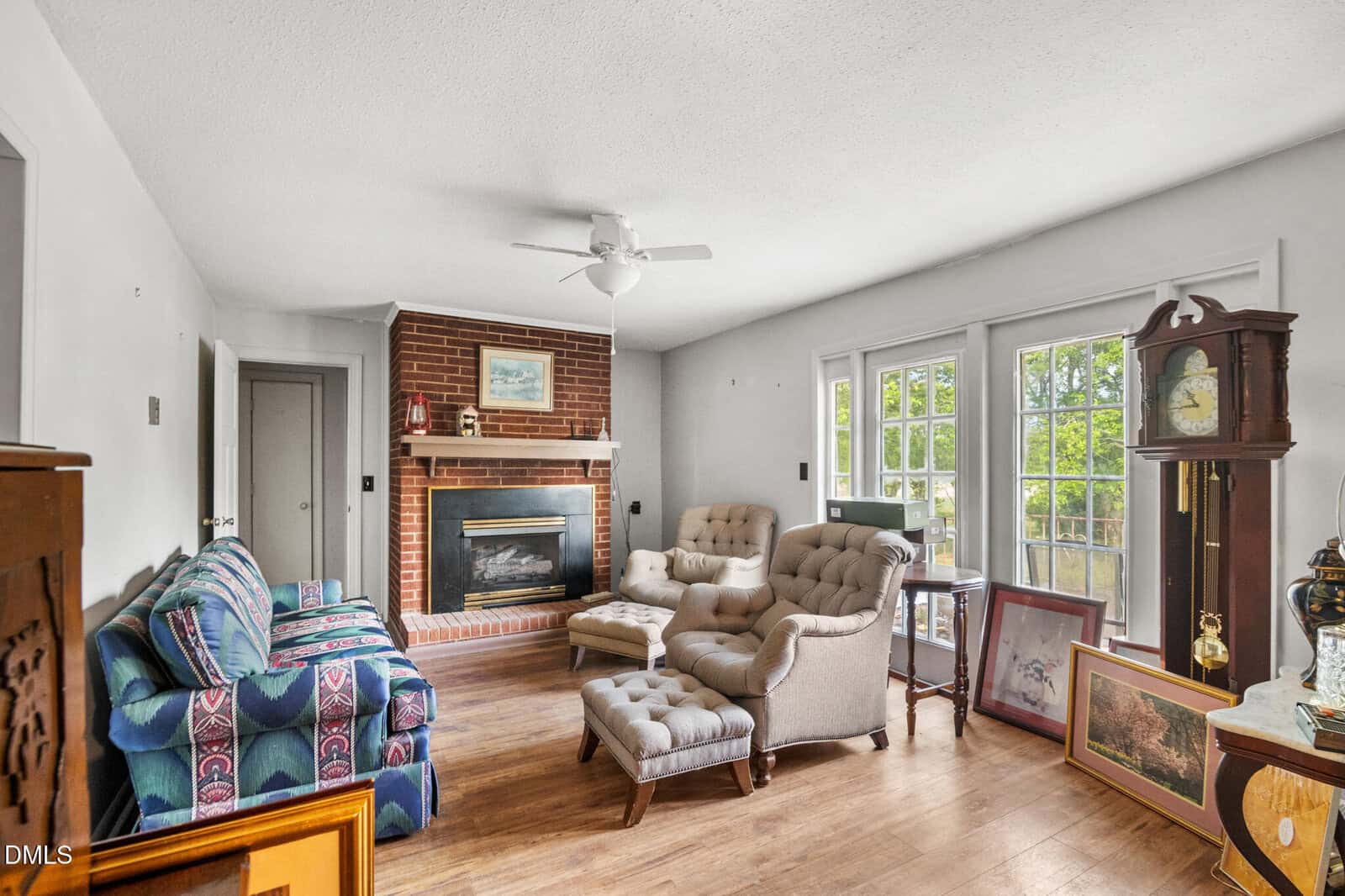 A cozy living room with a brick fireplace, ceiling fan, patterned sofa, tufted chairs, wooden floor, grandfather clock, and artwork leaning against glass doors with a view outside.