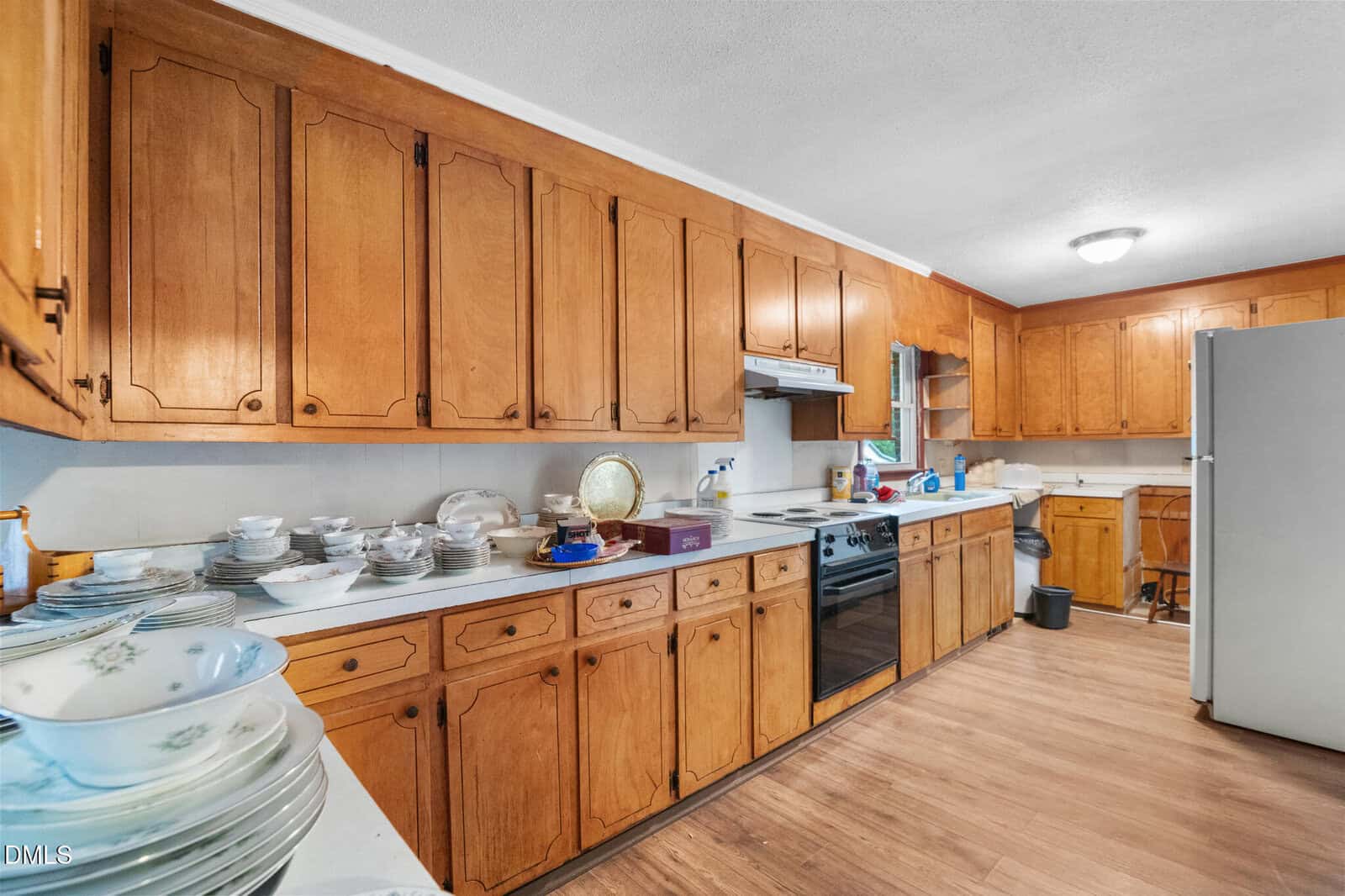 A kitchen with wooden cabinets, white countertops, an electric stove, a white refrigerator, and dishes stacked on the counters. The floor is wood, and there is a small window above the sink.