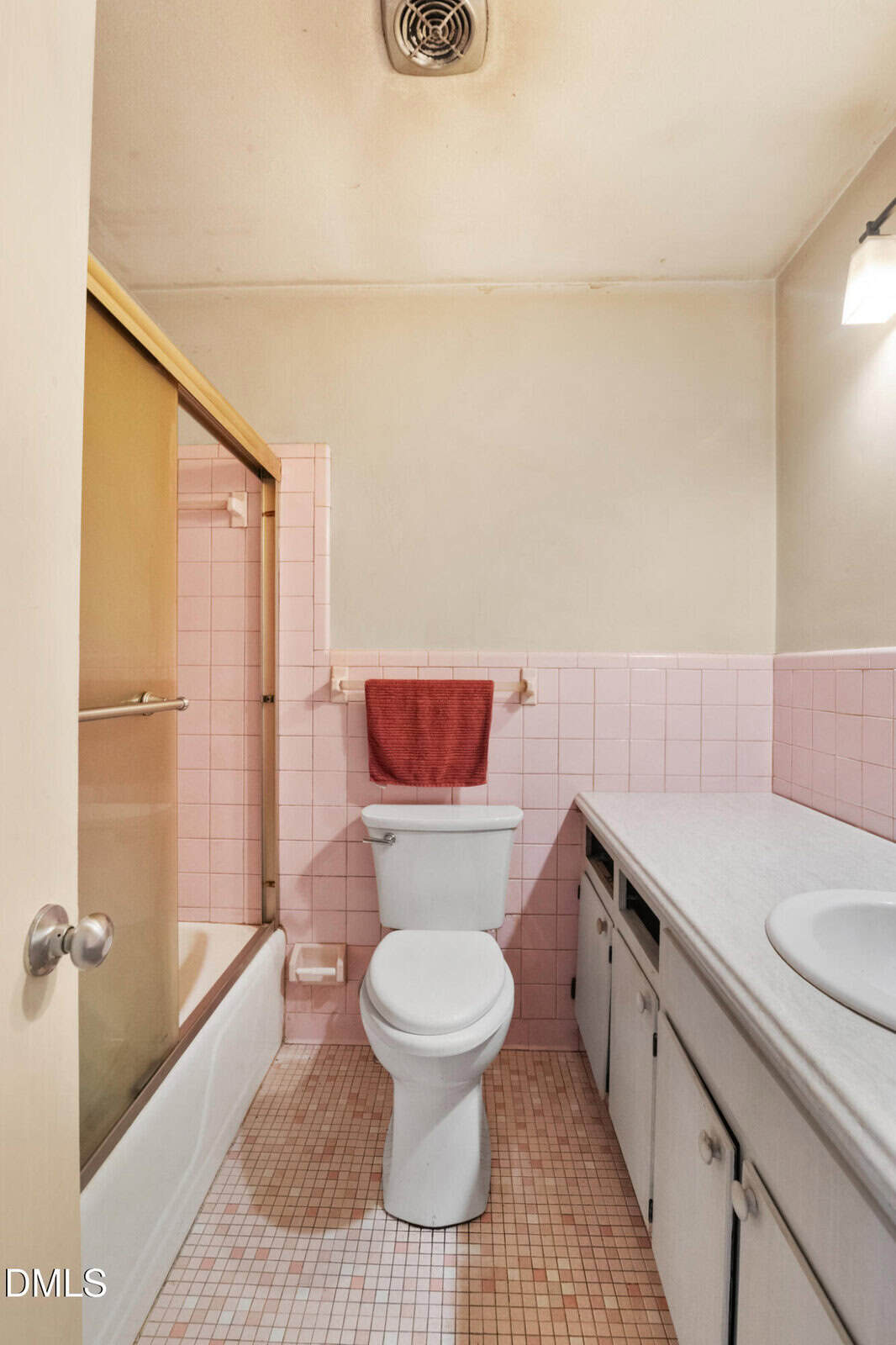 A small bathroom with a toilet, pink tile walls and floor, a bathtub with a sliding glass door, a white countertop, cabinets, and a red towel hanging behind the toilet.