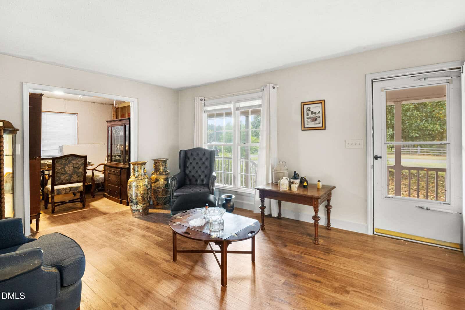 Bright living room with wood flooring, armchairs, a coffee table, large vases, and a window. Adjacent dining area visible through an open doorway. Glass door leads to outdoor porch.