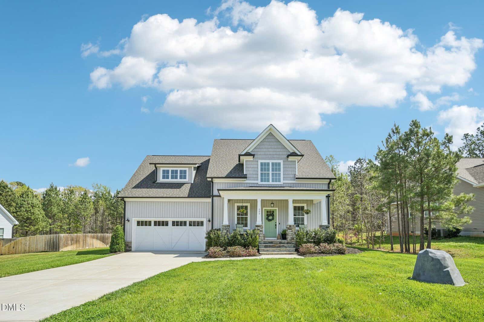 A modern two-story house with white siding, a covered front porch, and an attached two-car garage, surrounded by green grass, trees, and a blue sky with clouds.