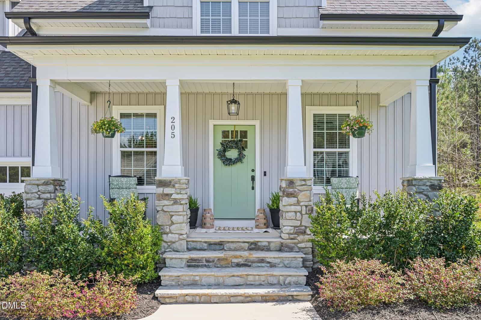 A modern house front with a mint green door, a wreath, stone steps, columns, potted plants, and white siding. Two windows frame the entrance, and there are shrubs and greenery along the walkway.