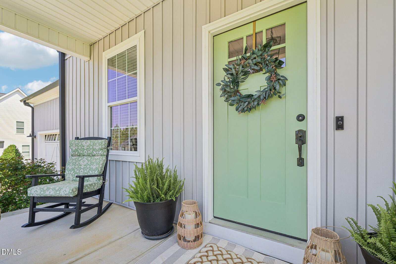 A light green front door with a wreath is on a covered porch, next to a window. A patterned rocking chair, potted plants, and woven baskets decorate the space. The house exterior is light gray siding.