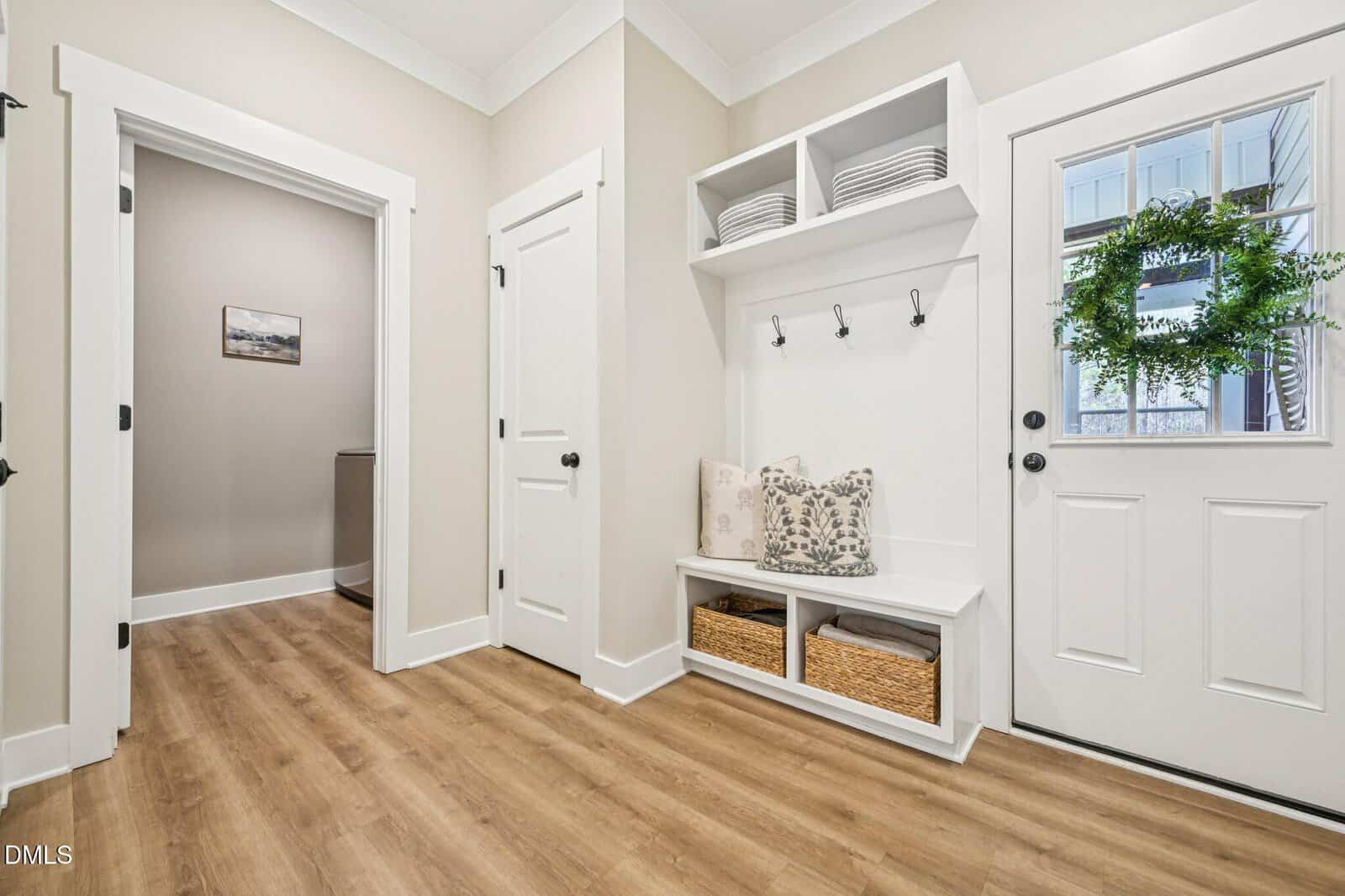 A bright mudroom with light wood flooring, white walls, a door with a wreath, built-in cubbies, hooks, two baskets, two pillows, and folded towels on shelves. An open doorway leads to another room.