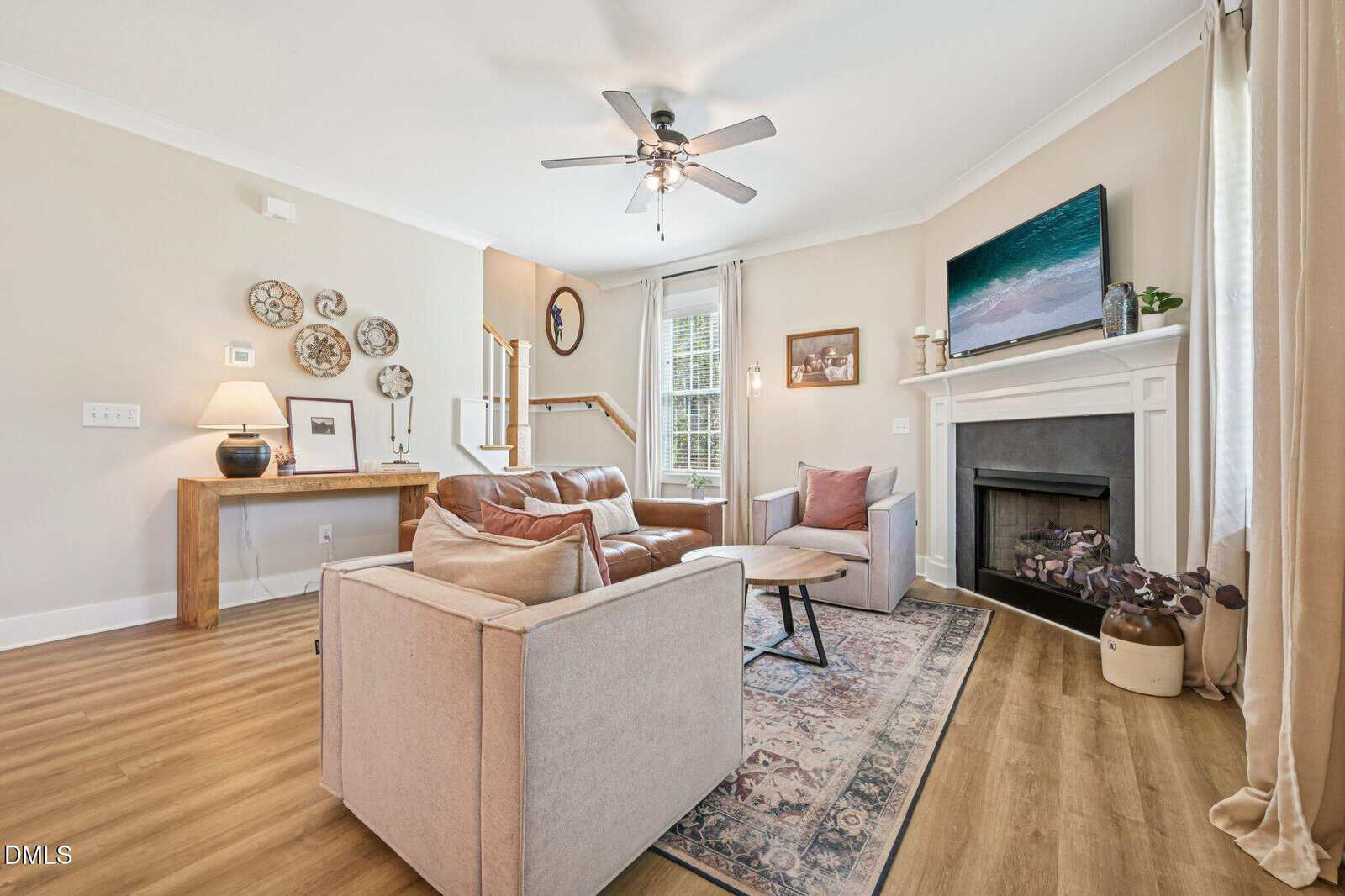 Cozy living room with a tan sofa, armchair, and rug, featuring a fireplace with a mounted TV above. Stairs and a table with decor are in the background, with light walls and wood floors brightened by natural light.