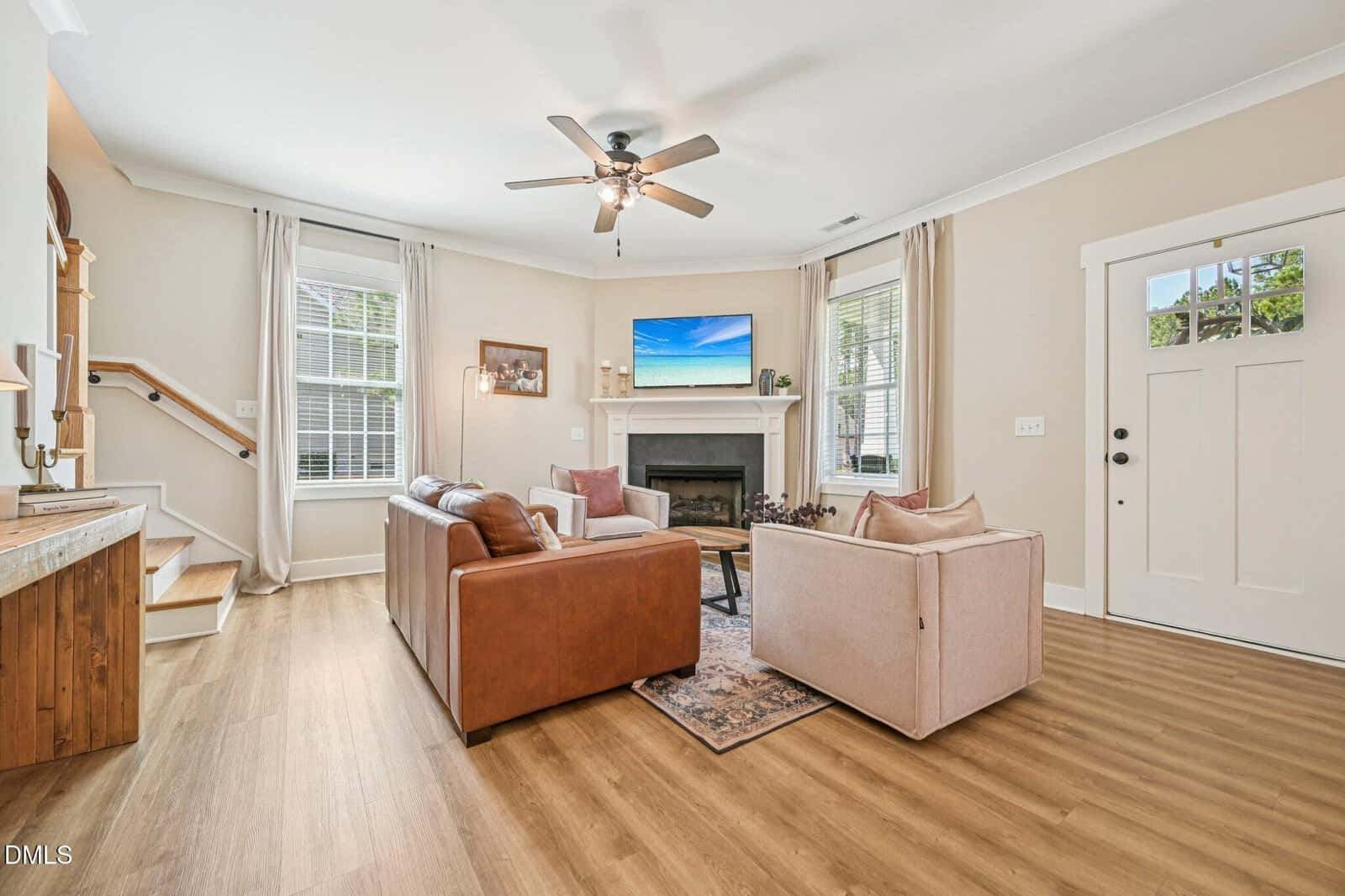 Bright living room with two sofas, a TV above a fireplace, large windows with curtains, wood flooring, a ceiling fan, and a staircase to the left. Door and natural light add a welcoming feel.