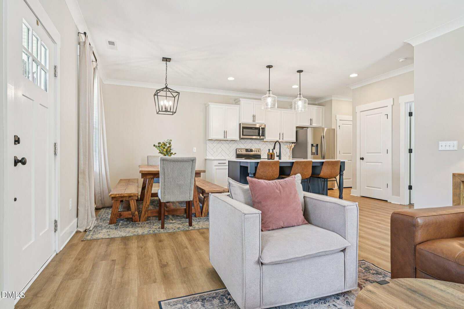 Modern open-concept living space with a gray armchair, pink pillow, wood dining table, and kitchen with white cabinets and stainless steel appliances. Light wood flooring and neutral decor throughout.
