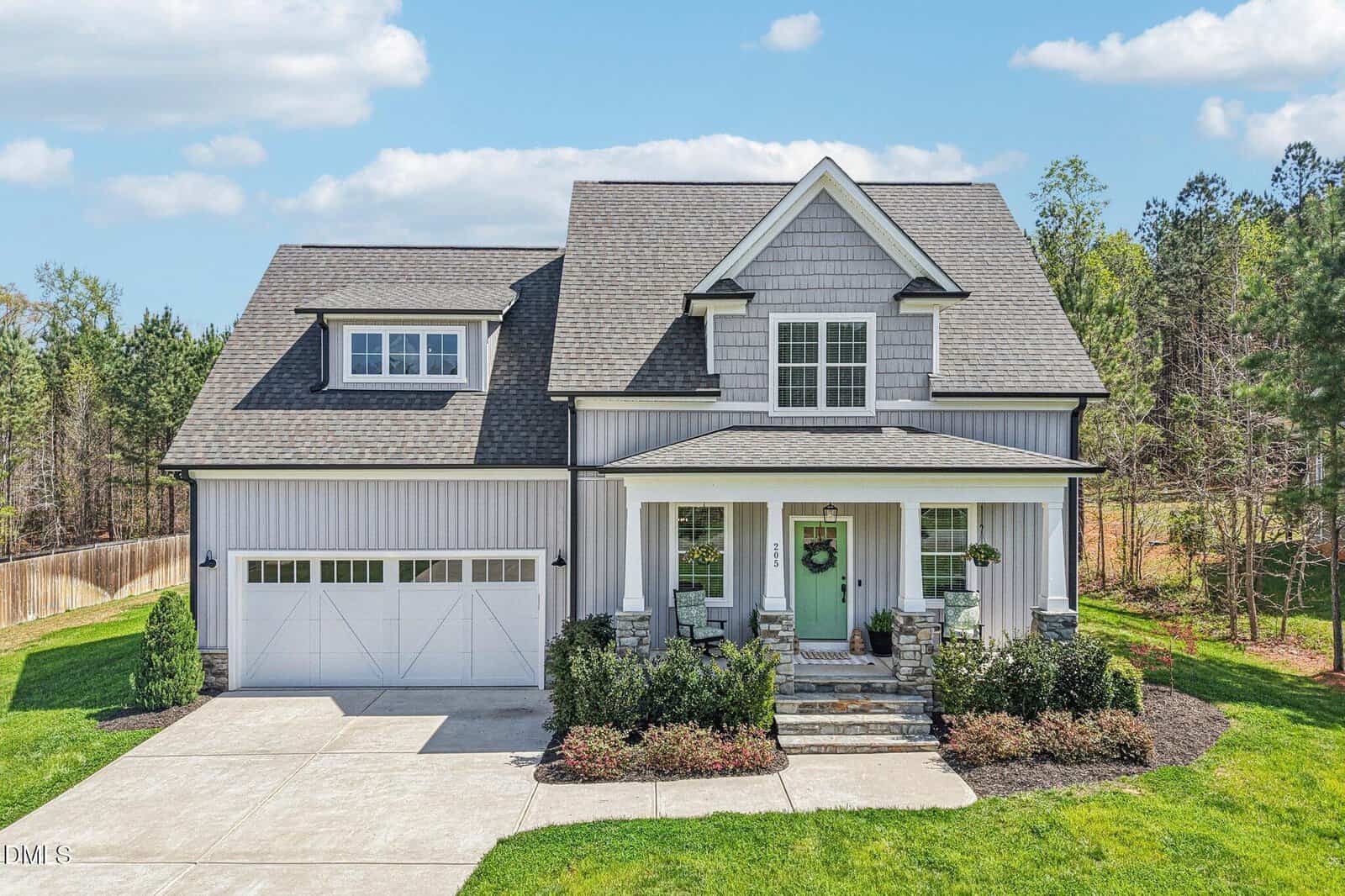 A modern two-story house with gray siding, a gable roof, double garage doors, and a covered front porch with stone columns, surrounded by green lawn, shrubs, and trees in the background.