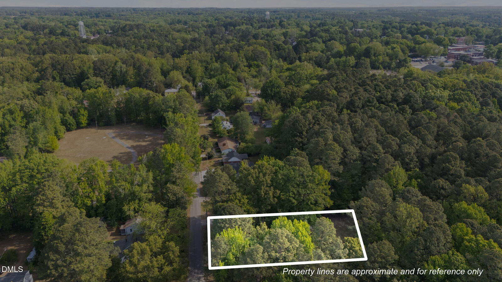 Aerial view of a wooded neighborhood with a rectangular plot of land outlined in white, indicating property boundaries. Surrounding areas are filled with dense trees and several houses.