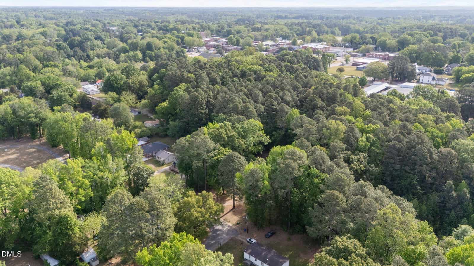 Aerial view of a neighborhood with houses surrounded by dense green trees, with a small town and open spaces visible in the background under a clear sky.