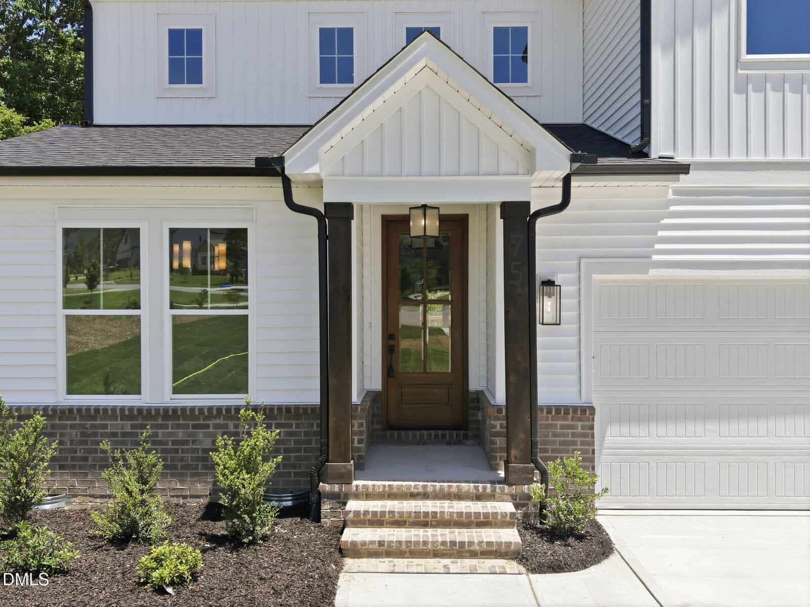 A modern house exterior with white siding, brick accents, and a wooden front door with glass panels. Small bushes are planted along the walkway, and a lantern-style light hangs above the entryway steps.
