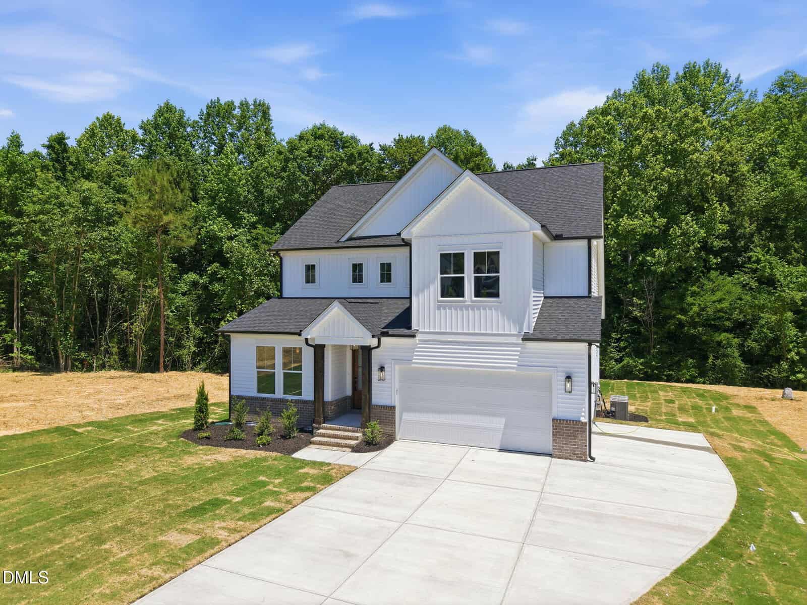 A modern two-story white house with black trim, a double garage, and a large driveway, set against a backdrop of green trees and a partly cloudy sky. Freshly laid grass is visible in the front yard.