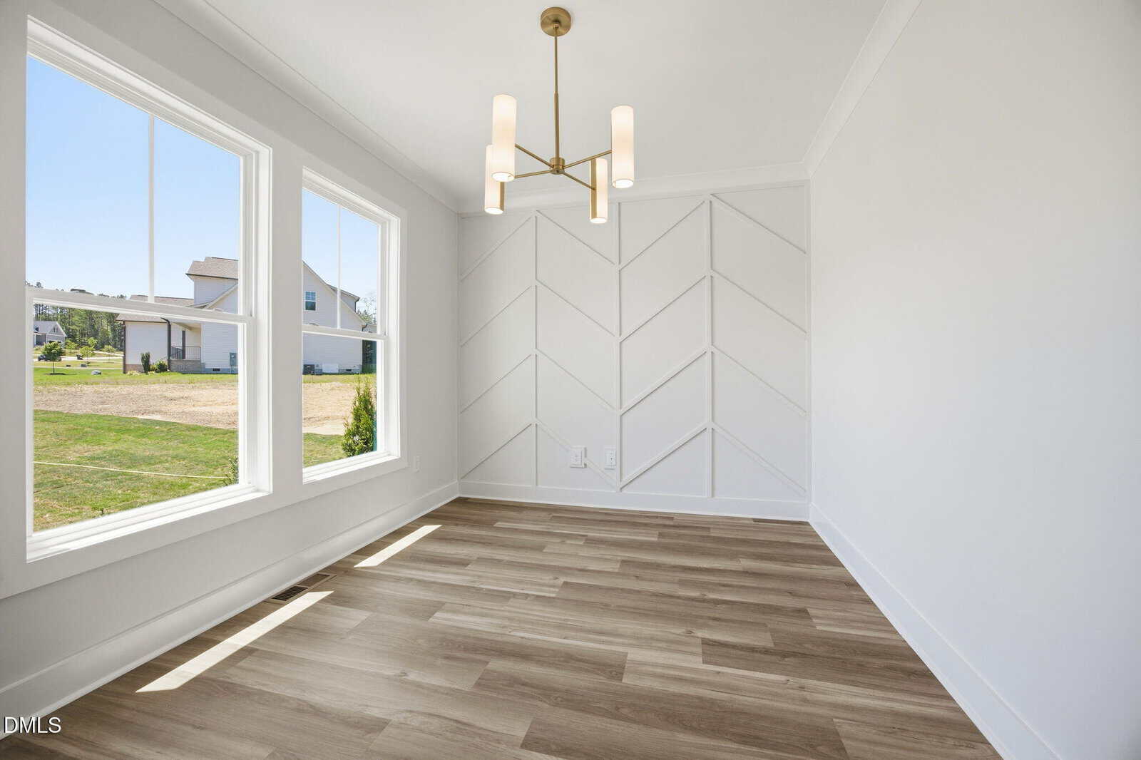 A bright, empty room with three large windows, light wood flooring, a white chevron accent wall, and a modern gold chandelier. The space is well-lit with natural light and has outdoor views.