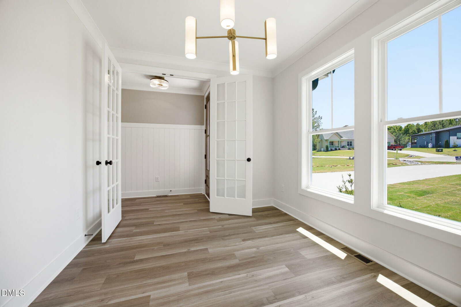 Bright, empty room with light wood flooring, large windows, modern ceiling light, white walls, and double glass French doors. The view outside shows grass, sidewalk, and houses in a residential area.