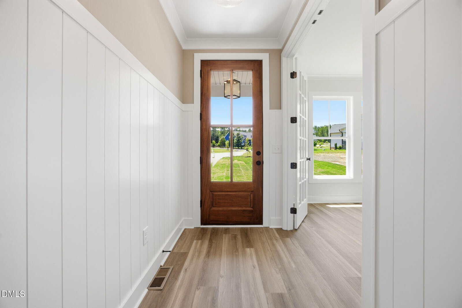Bright entryway with white paneled walls, light wood flooring, and a wooden front door with glass panes. A sunlit room with large windows is visible to the right, offering a view of green grass outside.