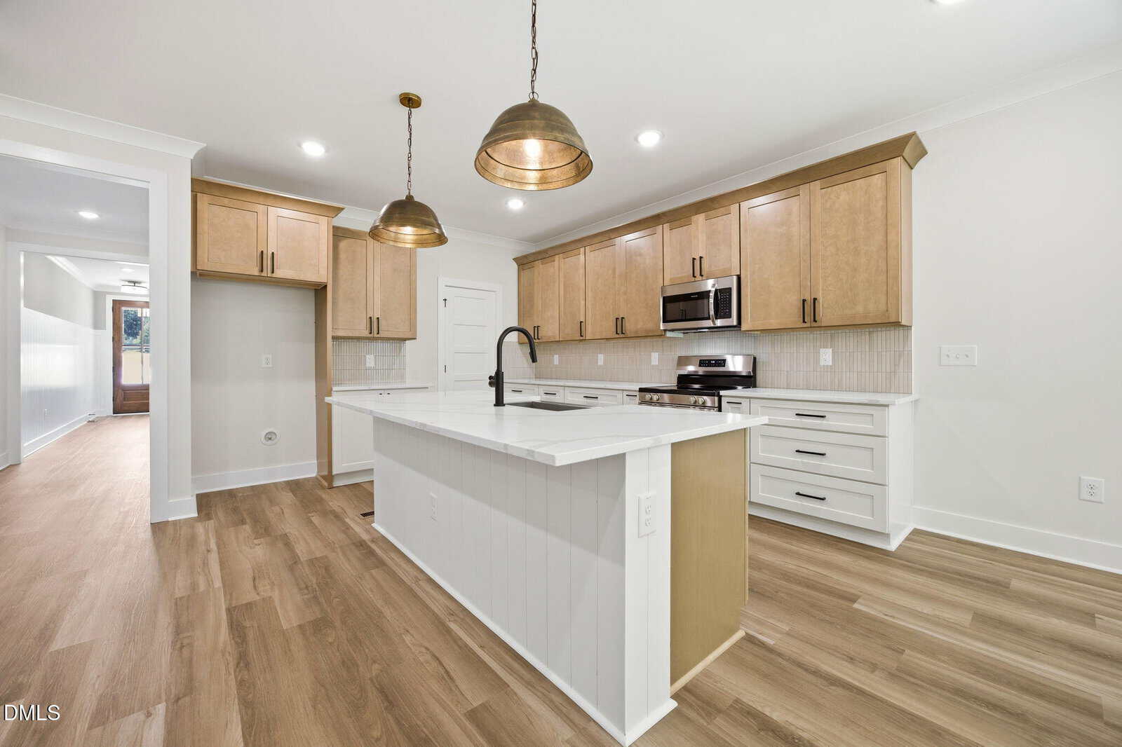 Modern kitchen with light wood cabinets, white countertops, stainless steel appliances, island with sink, two pendant lights, and light wood flooring. A hallway is visible to the left, and recessed lighting is in the ceiling.