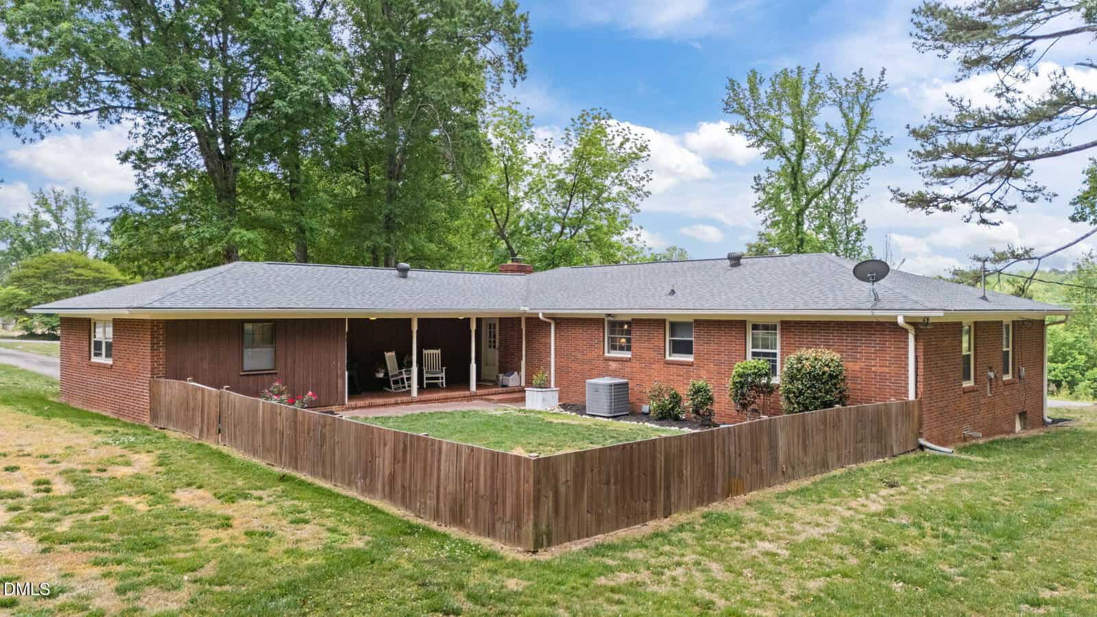 A one-story red brick house with a gray roof, wooden fenced patio, outdoor seating, and surrounded by green trees and grass under a blue sky with clouds.